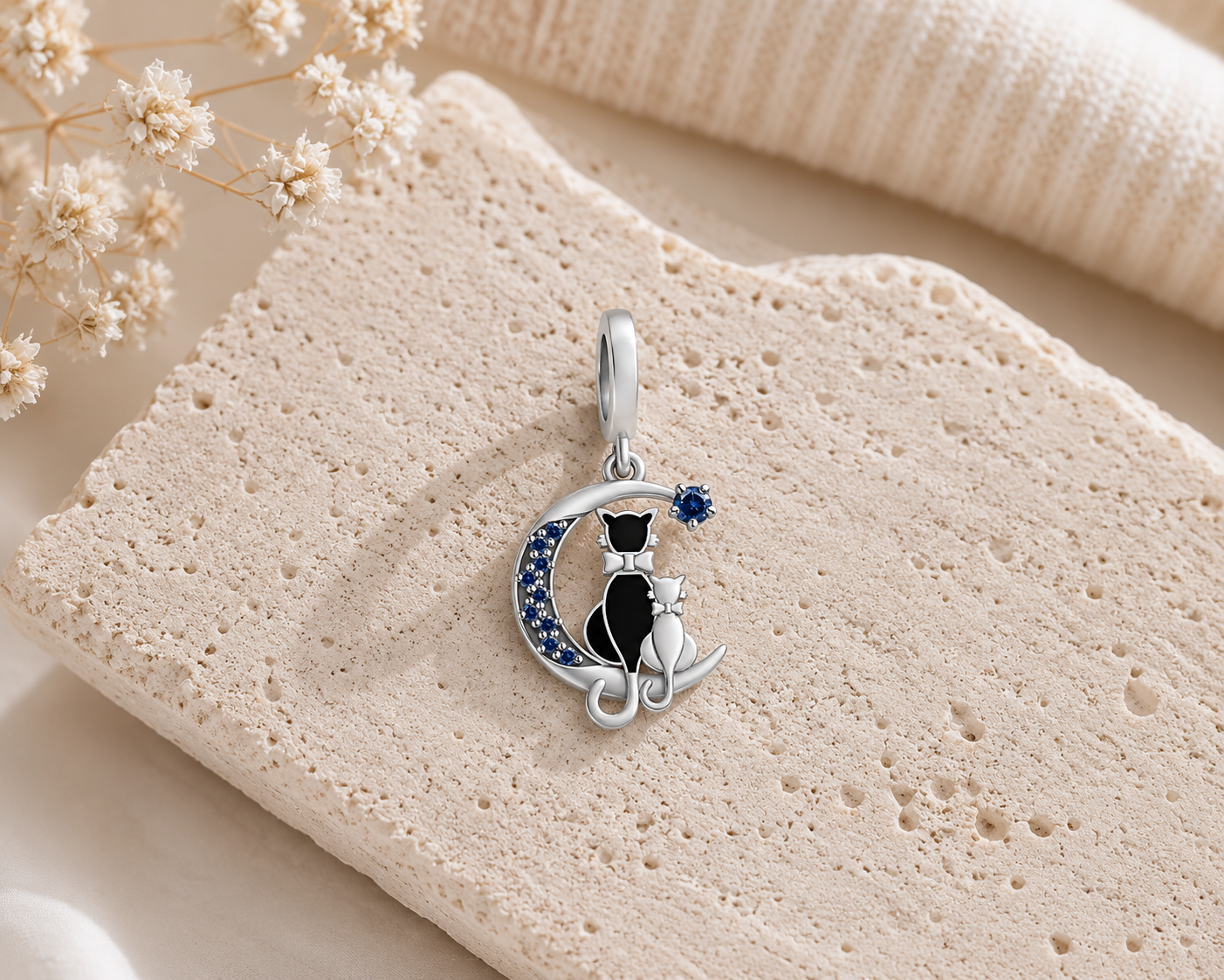 A silver pendant charm featuring a black cat sitting on a crescent moon accented with small blue stones, displayed on a textured beige stone slab with dried flowers.