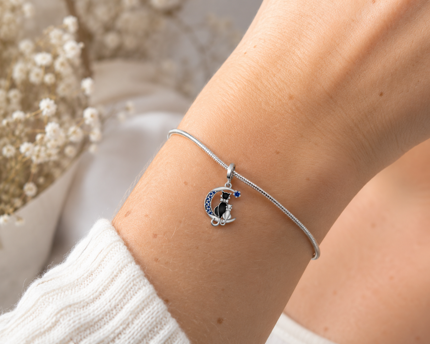 A close-up of a person's wrist wearing a delicate silver snake-braided bracelet with a charm depicting a black and white dog inside a crescent moon with blue star accents.