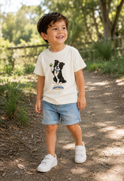 A young boy wearing a cream-colored graphic t-shirt with a black and white dog print, light blue denim shorts, and white sneakers, standing on a dirt path in a sunny outdoor setting.