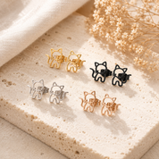 Cat-shaped earrings in gold, silver, and rose gold on a textured surface with dried flowers.
