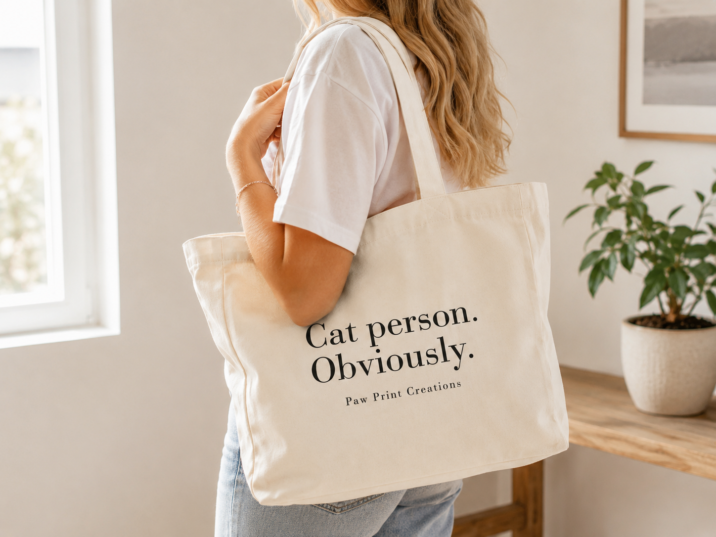 A woman in a white t-shirt and jeans carries a cream canvas tote bag with black text reading 'Cat person. Obviously.