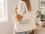 A woman in a white t-shirt and jeans carries a cream canvas tote bag with black text reading 'Cat person. Obviously.