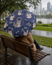 A navy blue umbrella with a repeating pattern of white cat portraits in oval frames is held over a seated person on a wooden park bench. The person is wearing a beige coat and dark pants, sitting in a city park with grass, water, and a skyline in the background.