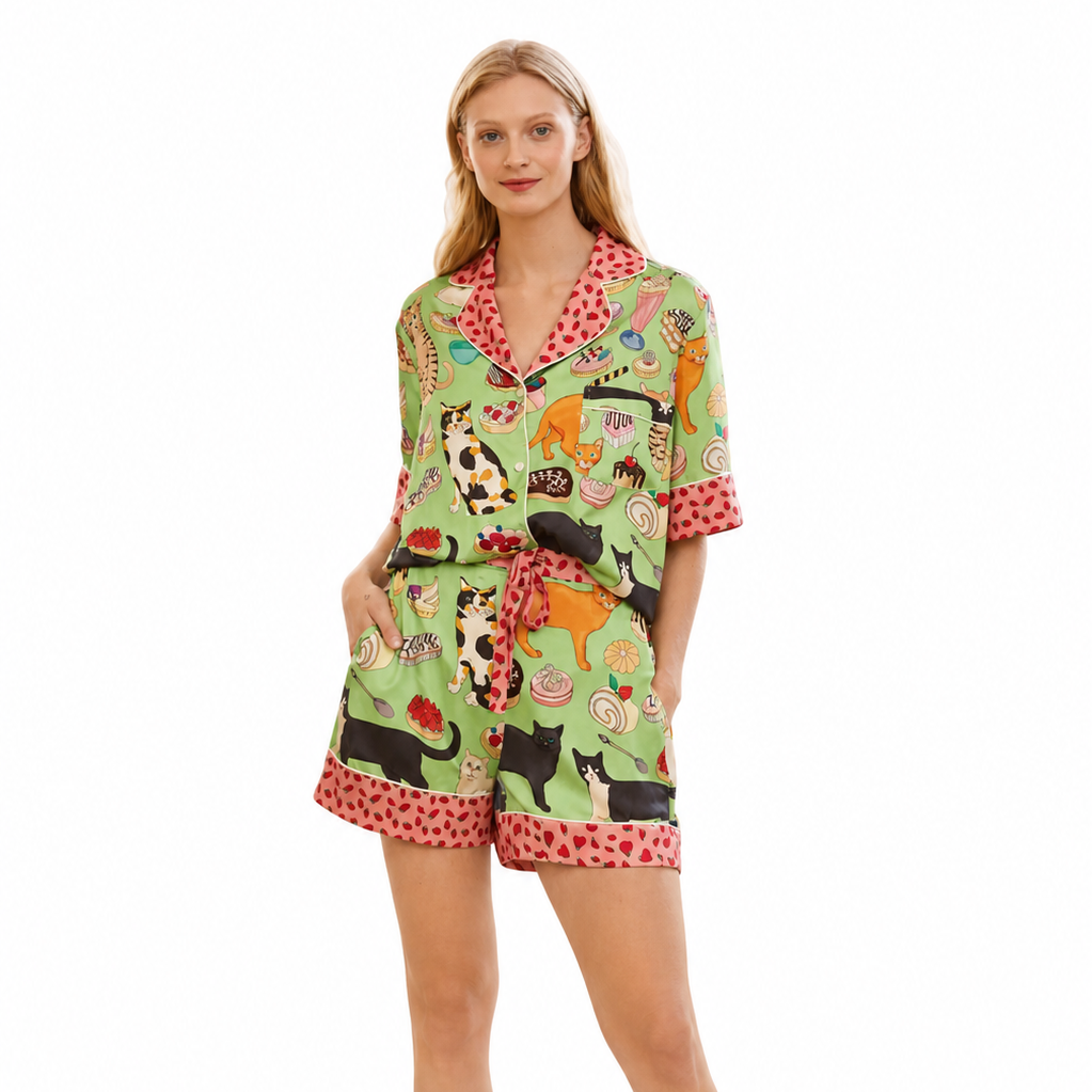 A woman modeling a matching two-piece pajama set with a colorful cartoon animal print on a light green background and red polka-dot trim, photographed against a plain white studio background.