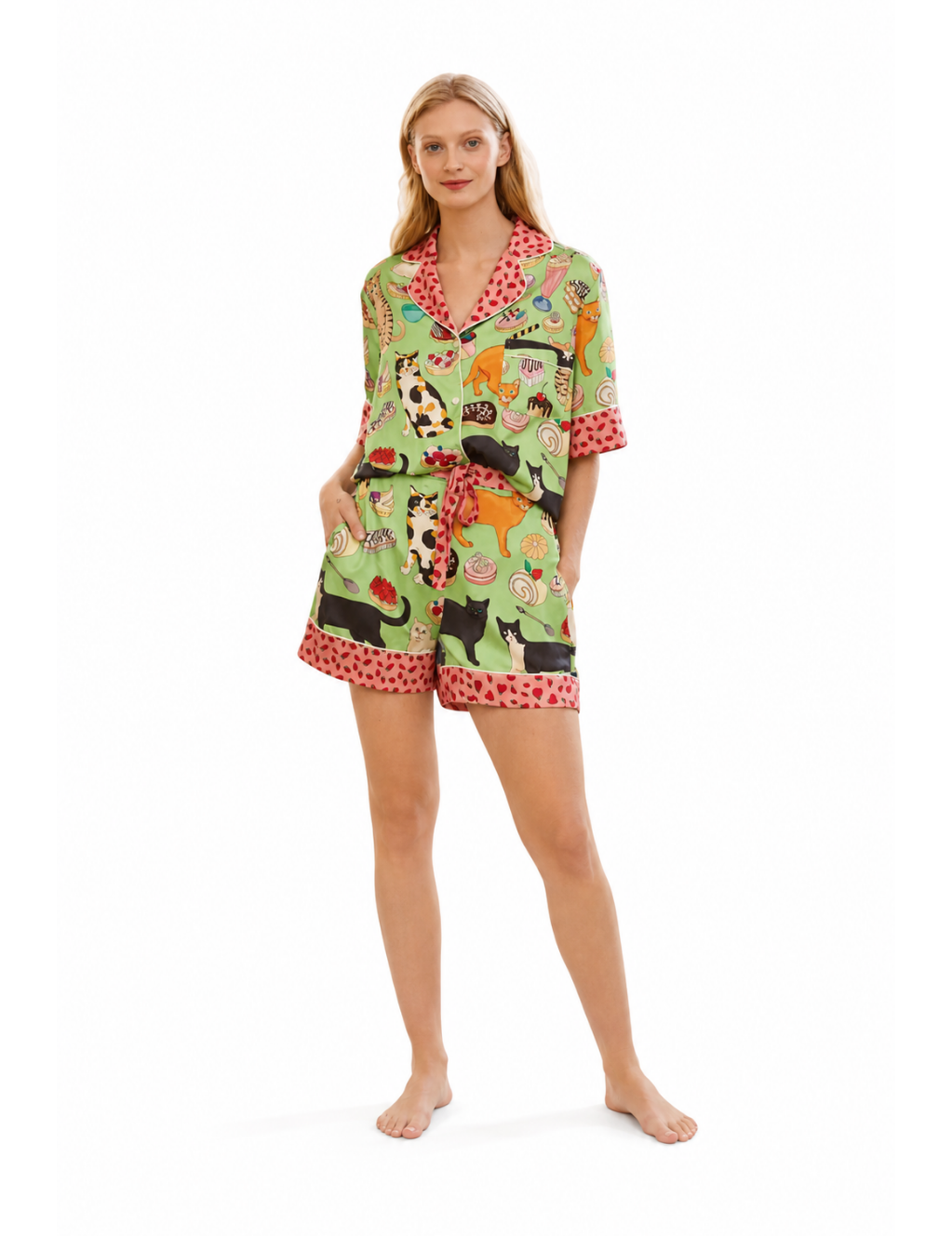 A woman modeling a matching two-piece pajama set with a colorful cartoon animal print on a light green background and red polka-dot trim, photographed against a plain white studio background.