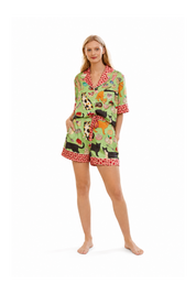 A woman modeling a matching two-piece pajama set with a colorful cartoon animal print on a light green background and red polka-dot trim, photographed against a plain white studio background.