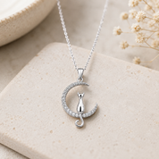 A silver necklace with a crescent moon pendant featuring a small cat silhouette and small clear stones, displayed on a beige stone surface with dried flowers in the background.