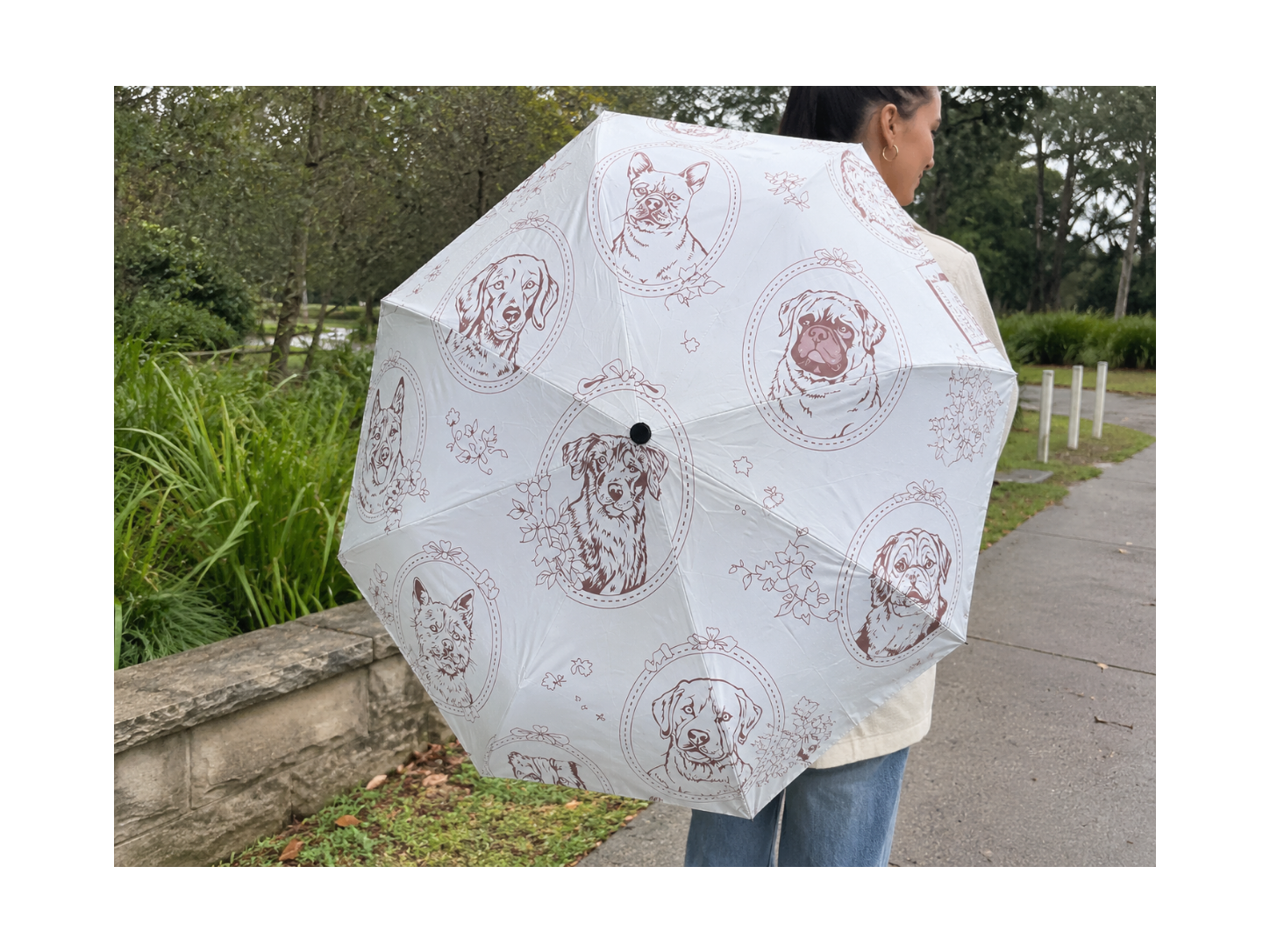 A person outdoors holding a white umbrella with a repeating brown line-art print of different dog breeds. The background shows a paved path and green foliage.