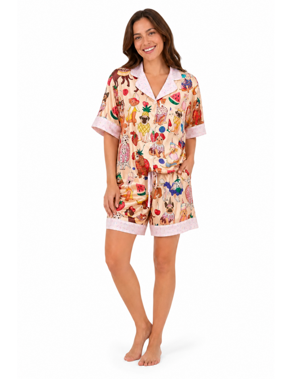 A woman modeling a two-piece pajama set featuring a colorful fruit and vegetable print on a light beige background, with light pink trim on the collar, sleeve cuffs, and shorts.