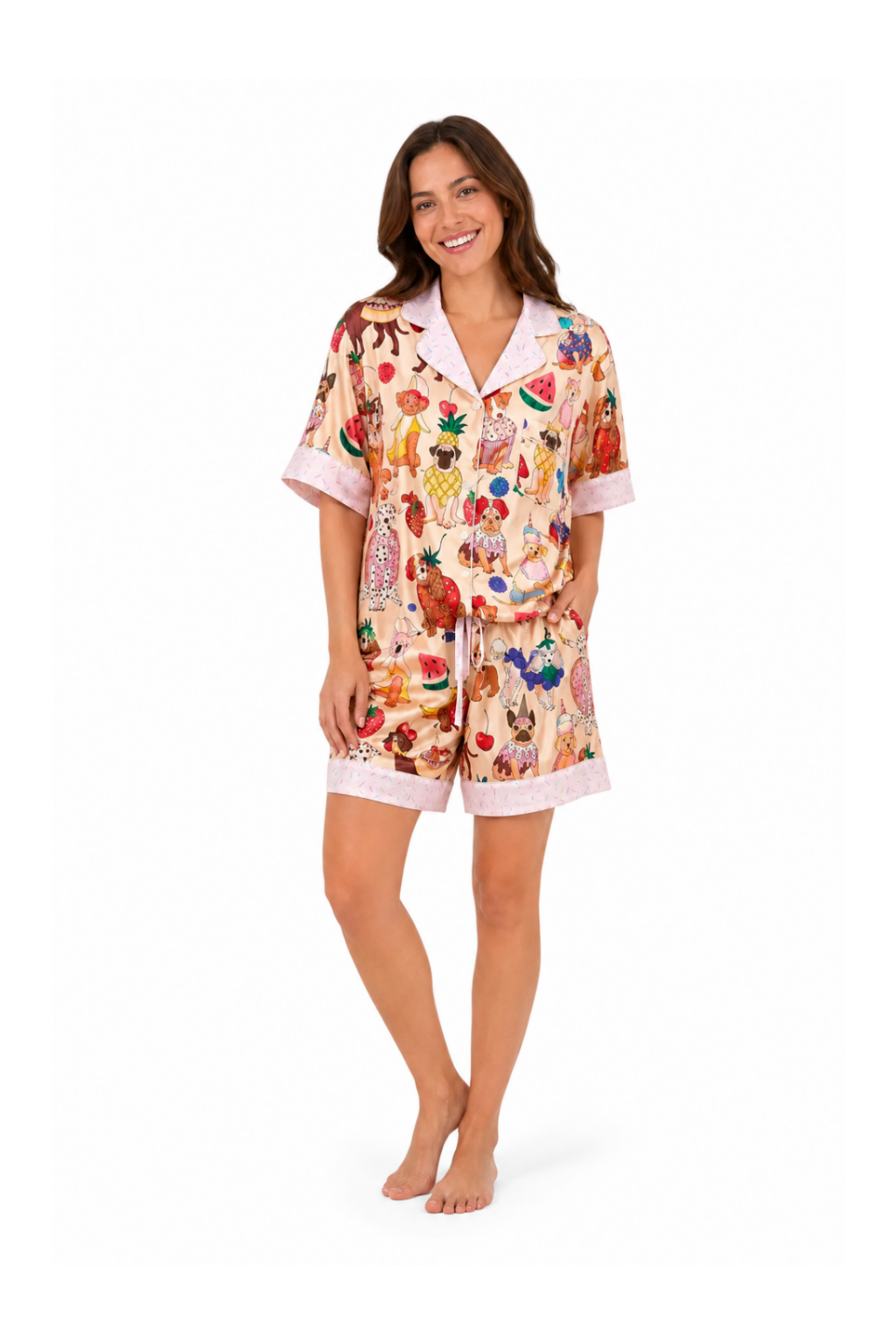 A woman modeling a two-piece pajama set featuring a colorful fruit and vegetable print on a light beige background, with light pink trim on the collar, sleeve cuffs, and shorts.