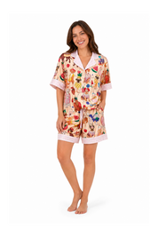 A woman modeling a two-piece pajama set featuring a colorful fruit and vegetable print on a light beige background, with light pink trim on the collar, sleeve cuffs, and shorts.