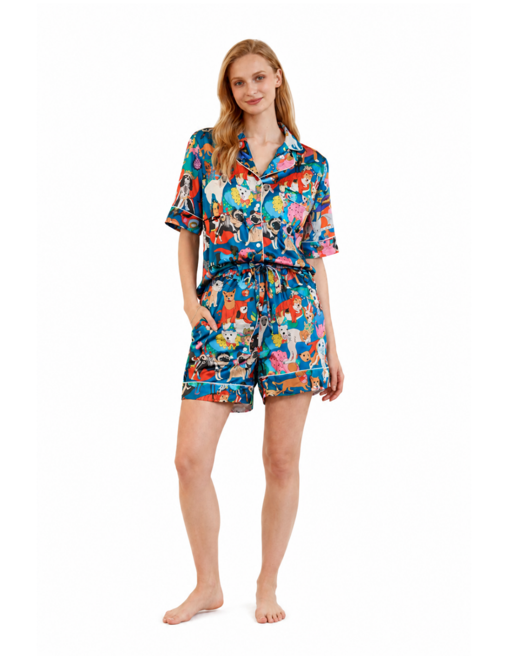A woman modeling a short-sleeve button-front pajama shirt and matching shorts set with a colorful cartoon print, photographed against a plain white studio background.