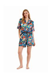 A woman modeling a short-sleeve button-front pajama shirt and matching shorts set with a colorful cartoon print, photographed against a plain white studio background.