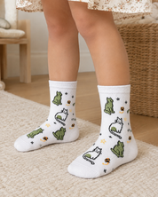 White crew socks with a playful pattern of cartoon frogs, cats, and small flowers, shown on a child's feet in a cozy indoor setting.