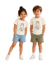 A young girl with a high ponytail stands against a white background wearing a cream-colored t-shirt with a cartoon cat graphic and the text 'Whisker Friends', paired with denim shorts and white sneakers.