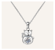 A silver-toned necklace featuring a cat-shaped pendant with a raised paw and a clear round gemstone set on its belly, shown against a light beige background.