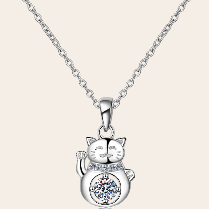 A silver-toned necklace featuring a cat-shaped pendant with a raised paw and a clear round gemstone set on its belly, shown against a light beige background.