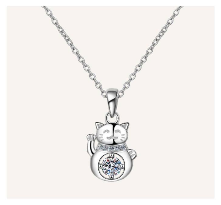 A silver-toned necklace featuring a cat-shaped pendant with a raised paw and a clear round gemstone set on its belly, shown against a light beige background.