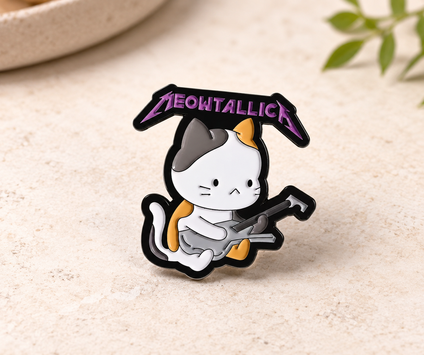 A metal pin featuring a cartoon calico cat playing a guitar under a purple 'MEOWTALLICA' banner, displayed on a textured beige surface.