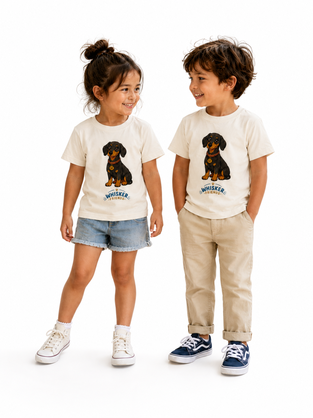 A young child standing against a white studio background, wearing a cream short-sleeve T-shirt with a dachshund graphic and the word 'WHISKER' below it, paired with blue denim shorts and white shoes.
