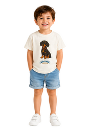 A young child standing against a white studio background, wearing a cream short-sleeve T-shirt with a dachshund graphic and the word 'WHISKER' below it, paired with blue denim shorts and white shoes.