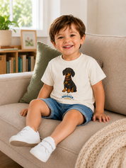 A young boy sitting on a beige sofa wearing a white t-shirt with a black and tan dachshund graphic and the text 'Whisker & Co'.