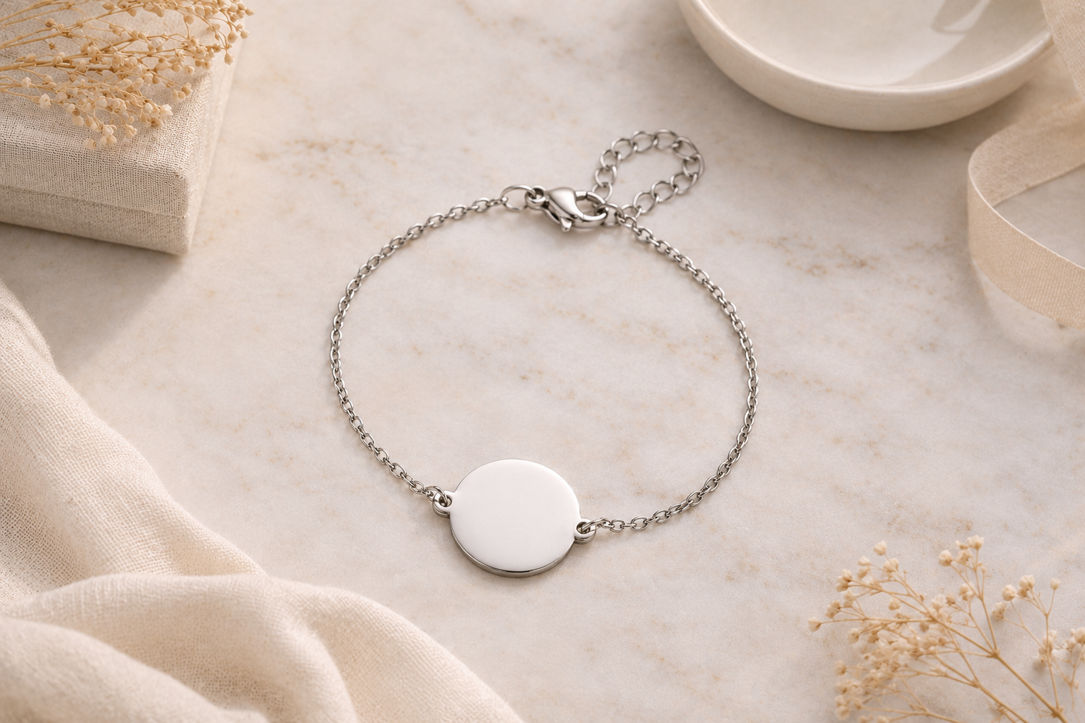 A delicate silver chain bracelet with a round, blank white disc charm, styled on a light marble surface with soft beige fabric and dried flowers.