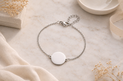 A delicate silver chain bracelet with a round, blank white disc charm, styled on a light marble surface with soft beige fabric and dried flowers.