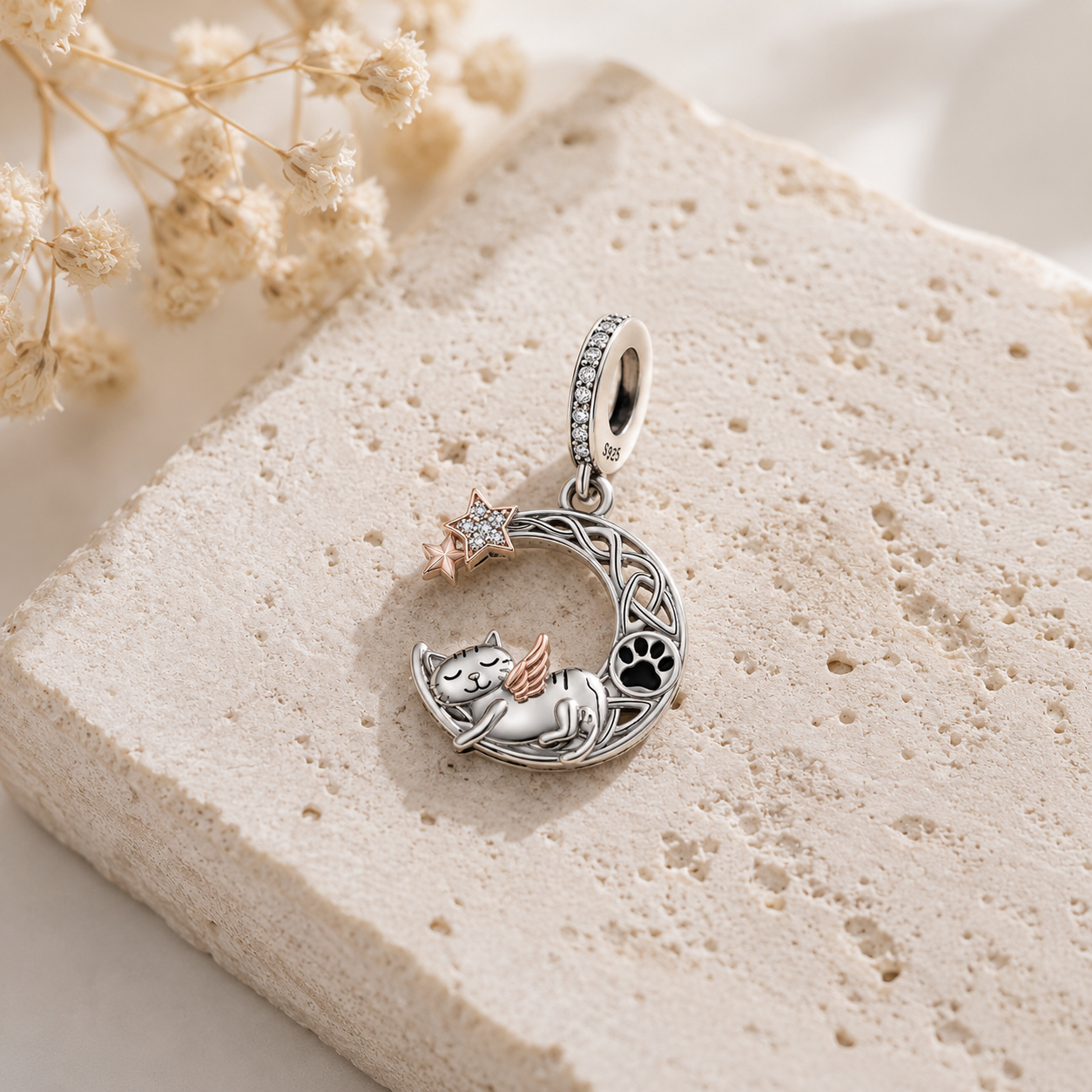 A silver crescent moon pendant charm featuring a sleeping cat, a paw print, and a small rose-gold star, displayed on a textured beige stone slab with dried flowers.
