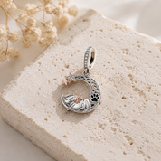 A silver crescent moon pendant charm featuring a sleeping cat, a paw print, and a small rose-gold star, displayed on a textured beige stone slab with dried flowers.