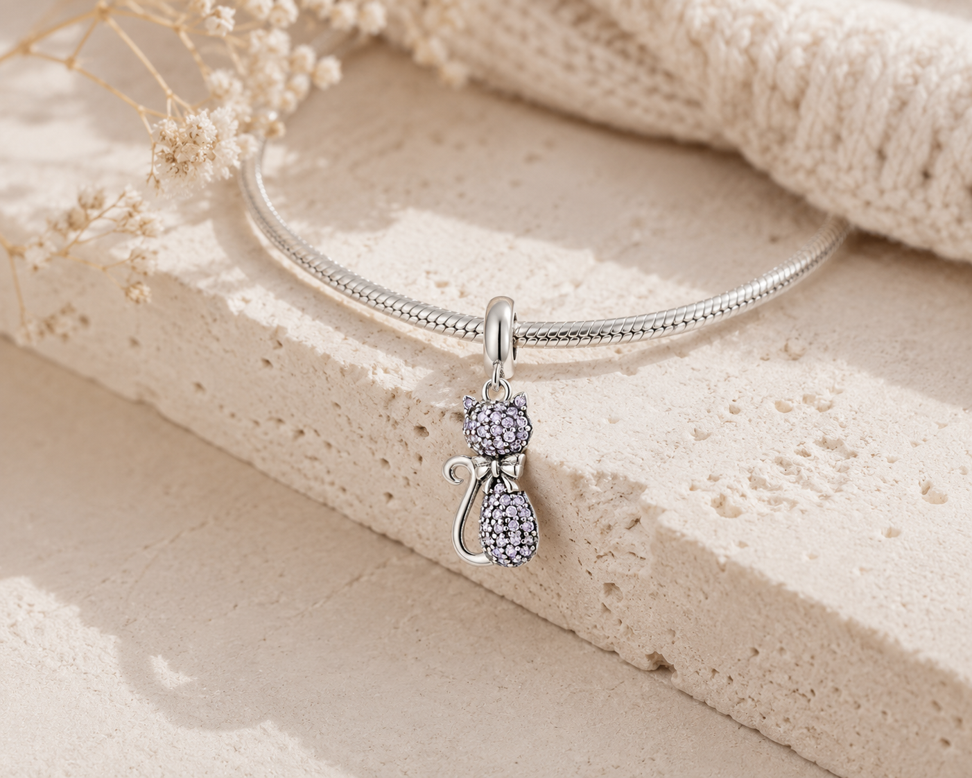 A silver snake chain bracelet with a dangling cat charm encrusted with clear rhinestones, styled on a textured beige stone slab with dried flowers and a cream knit fabric.