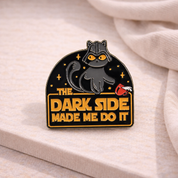 A black enamel pin featuring a cartoon cat with yellow eyes and the text 'THE DARK SIDE MADE ME DO IT' in yellow, displayed on a light beige fabric surface.