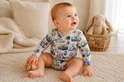 A baby sitting on a cream quilted bed wearing a long-sleeve cream bodysuit with a blue toile pattern of cats and boats. The background features neutral-toned pillows and a plush bunny toy.