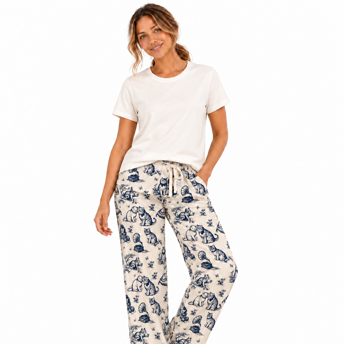 A person wearing cream-colored lounge pants with a blue toile-style print of cats, bowls, and leaves, shown from the waist down against a light gray studio background.