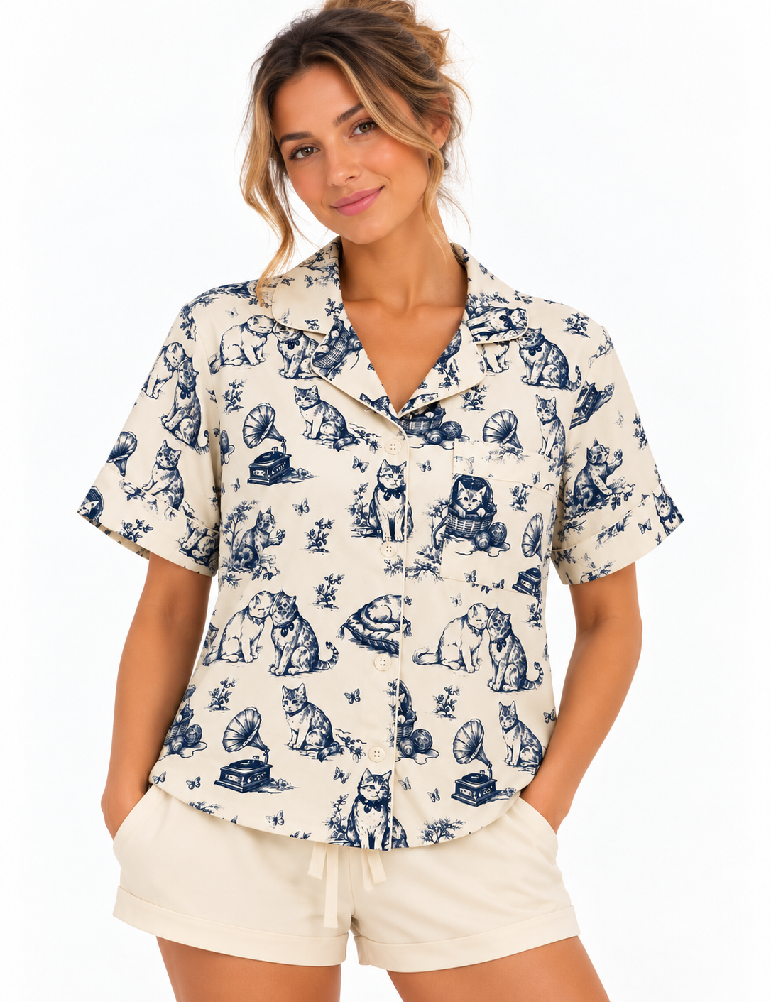 A woman wearing a short-sleeve button-front pajama shirt with a blue toile-style print of cats and dogs on a warm off-white background, photographed against a clean white studio background.