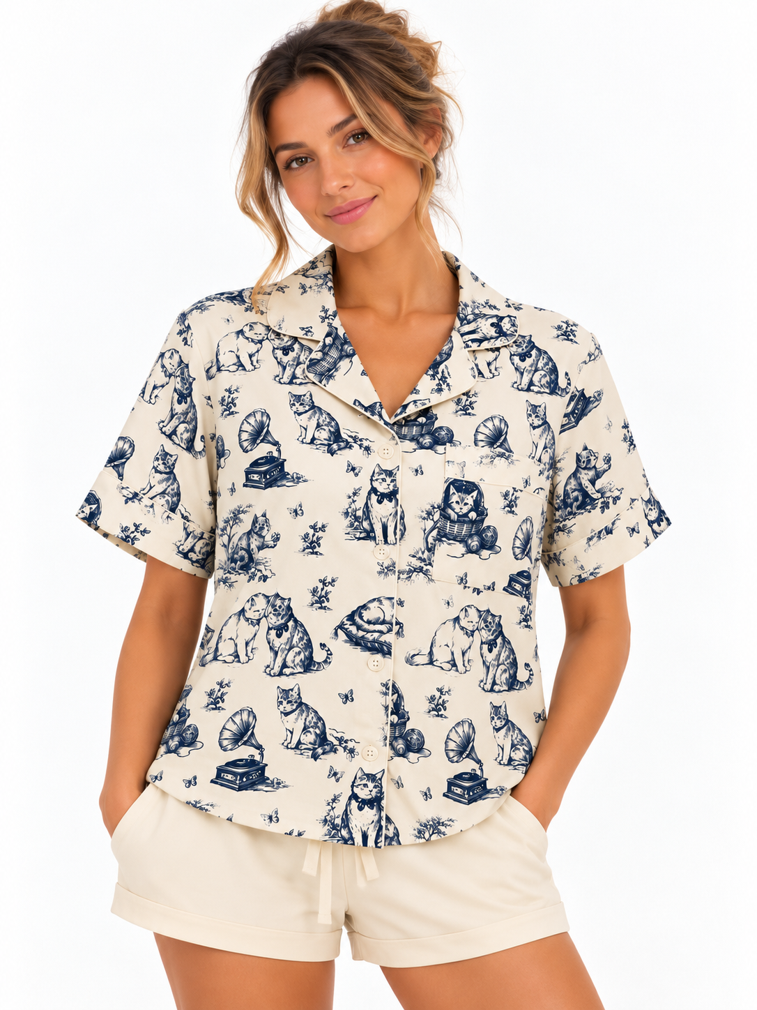 A woman wearing a short-sleeve button-front pajama shirt with a blue toile-style print of cats and dogs on a warm off-white background, photographed against a clean white studio background.