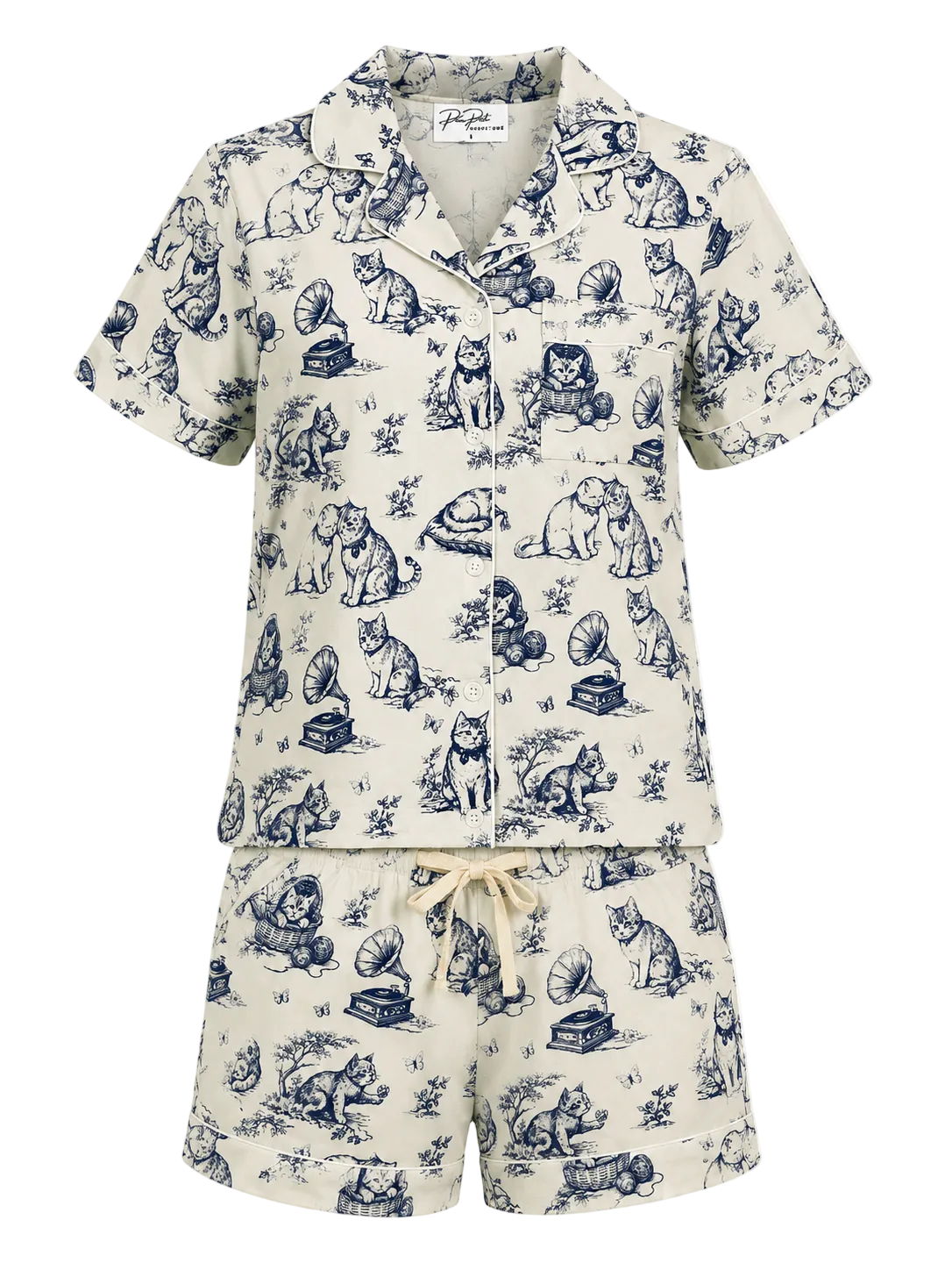 A two-piece short-sleeve pajama set featuring a matching button-up top and shorts in cream with a navy blue toile de Jouy print of dogs and cats, complete with a cream drawstring tie at the waist.