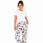 A woman modeling a matching short-sleeve button-up pajama shirt and drawstring pants set in an off-white fabric with a blue toile-style print of dogs and baskets, against a plain white background.