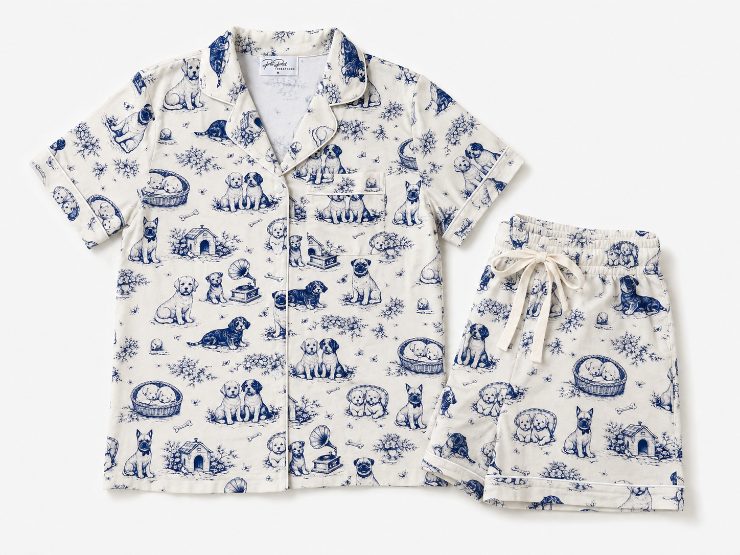 A two-piece pajama set featuring a short-sleeve button-front top and matching shorts in an off-white fabric with a blue toile-style dog pattern, laid flat on a light gray background.
