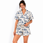 A woman modeling a matching short-sleeve button-front pajama top and shorts set with a blue toile-style print on an off-white background, photographed against a clean white studio backdrop.