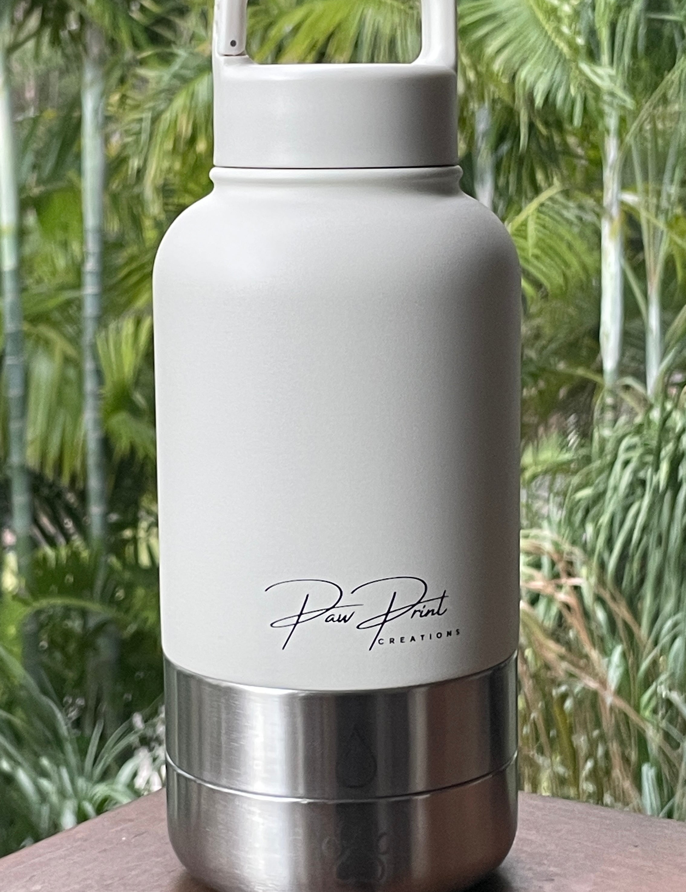 A white insulated water bottle with a stainless steel base and a white carrying loop lid, featuring the 'Paw Print Creations' brand logo, set on a wooden ledge with a blurred green tropical background.