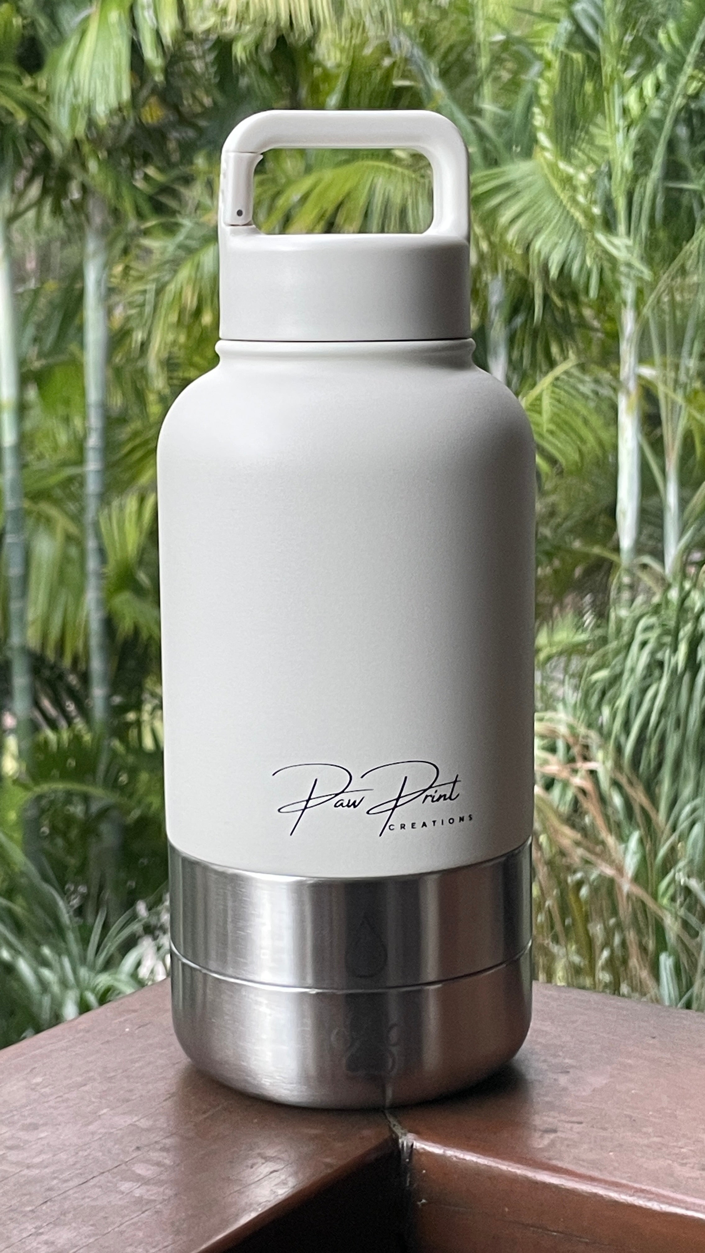 A white insulated water bottle with a stainless steel base and a white carrying loop lid, featuring the 'Paw Print Creations' brand logo, set on a wooden ledge with a blurred green tropical background.