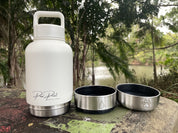 A white insulated water bottle with a stainless steel base and two detached stainless steel bowls placed beside it on a stone surface outdoors, with trees and water in the background. The bottle has a visible script logo reading 'Paw Print Creations'.