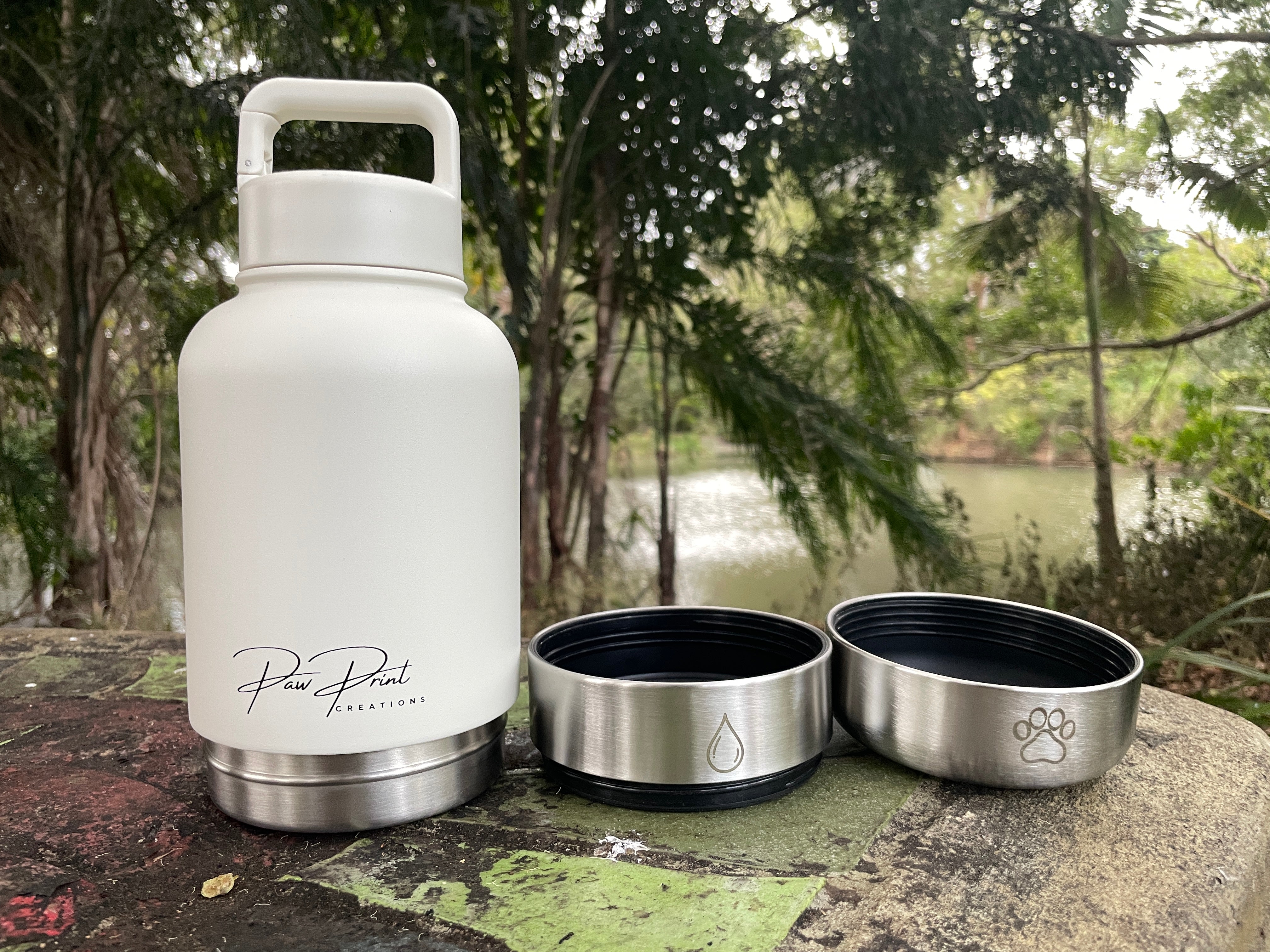 A white insulated water bottle with a stainless steel base and two detached stainless steel bowls placed beside it on a stone surface outdoors, with trees and water in the background. The bottle has a visible script logo reading 'Paw Print Creations'.