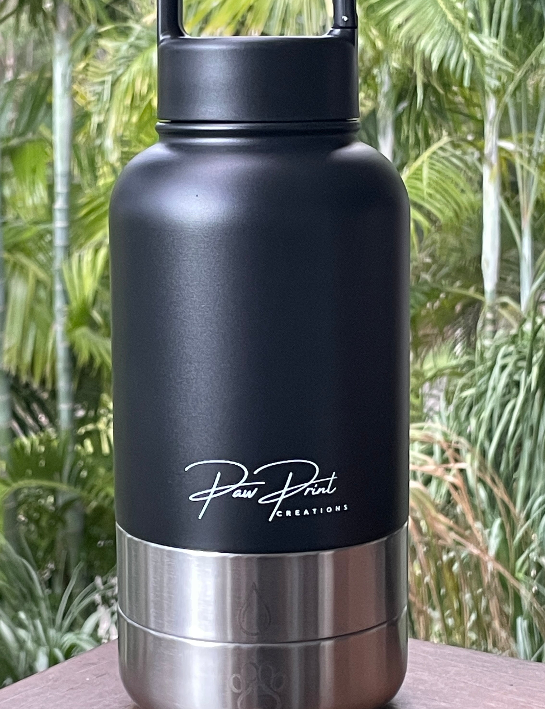 A matte black insulated water bottle with a stainless steel base and a carry handle, standing on a wooden surface with a blurred tropical green background. The bottle features the 'Paw Paw Creations' brand logo on the front.