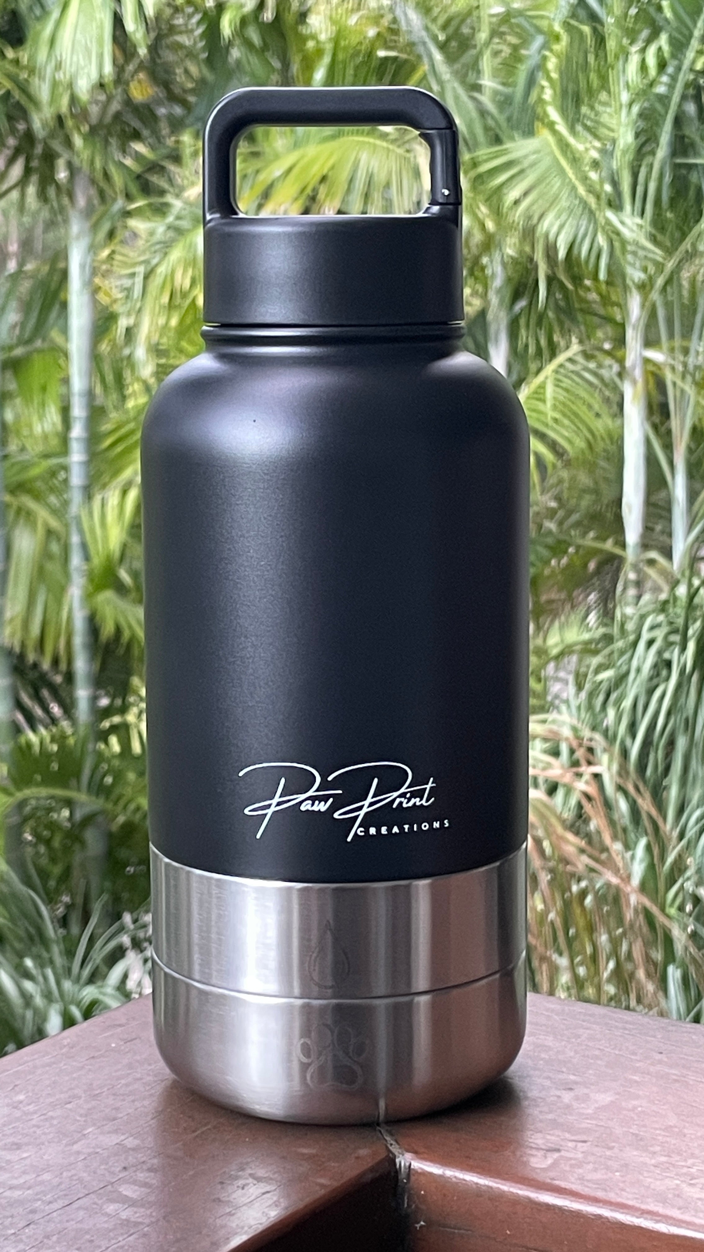 A matte black insulated water bottle with a stainless steel base and a carry handle, standing on a wooden surface with a blurred tropical green background. The bottle features the 'Paw Paw Creations' brand logo on the front.