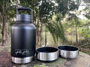 A matte black insulated water bottle with a carry loop and silver base, shown outdoors on a stone surface with two detached stainless steel lids beside it. Trees and a body of water are blurred in the background.