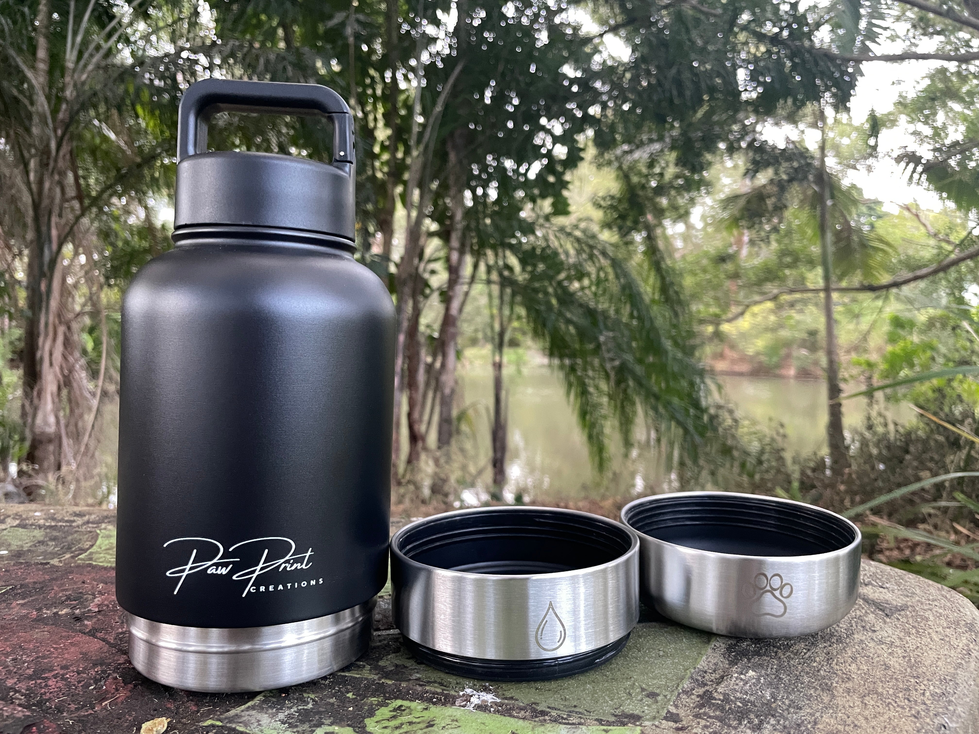 A matte black insulated water bottle with a carry loop and silver base, shown outdoors on a stone surface with two detached stainless steel lids beside it. Trees and a body of water are blurred in the background.
