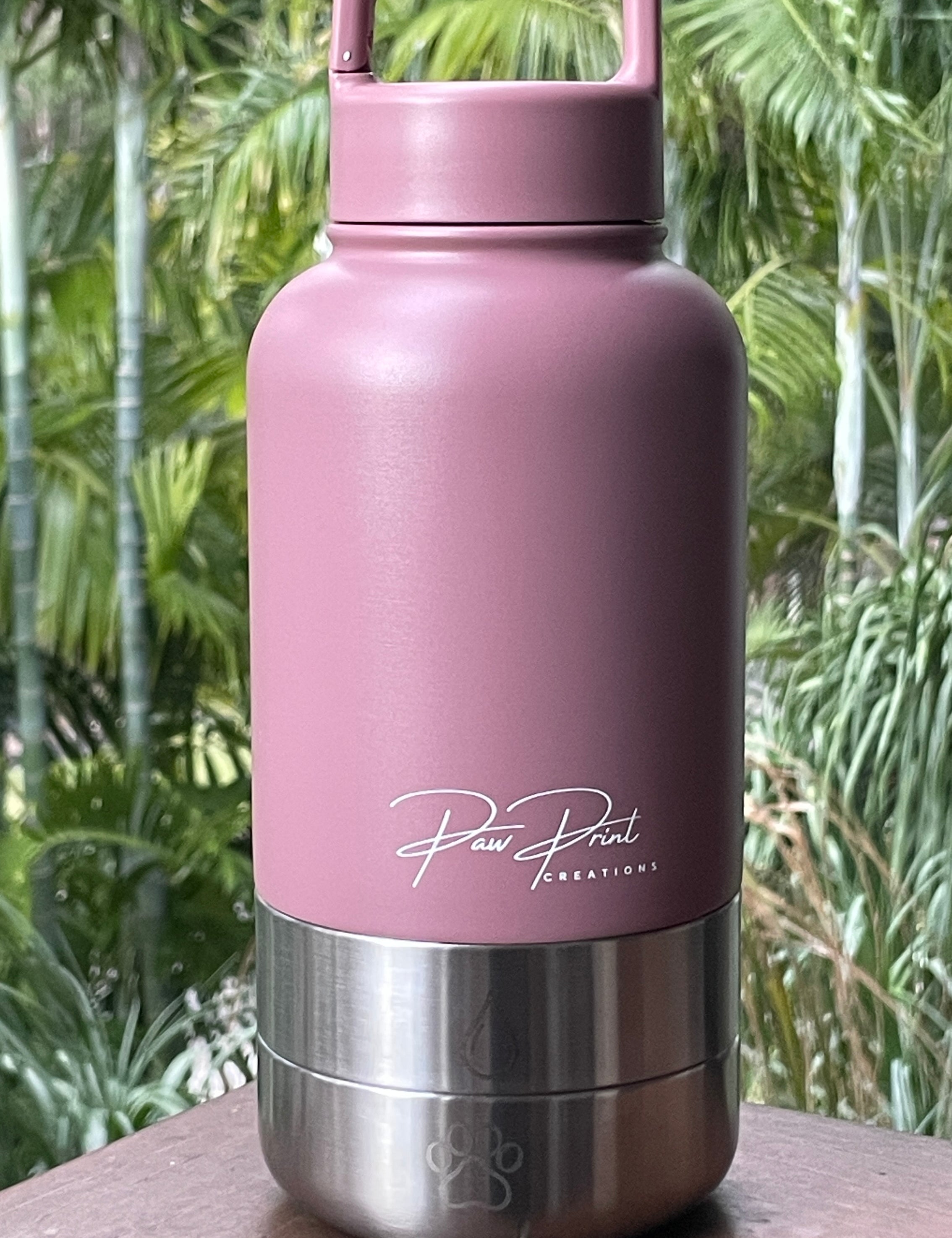 A dusty pink insulated water bottle with a stainless steel base, featuring the 'Paw Print Creations' logo, resting on a wooden ledge with a blurred tropical green background.