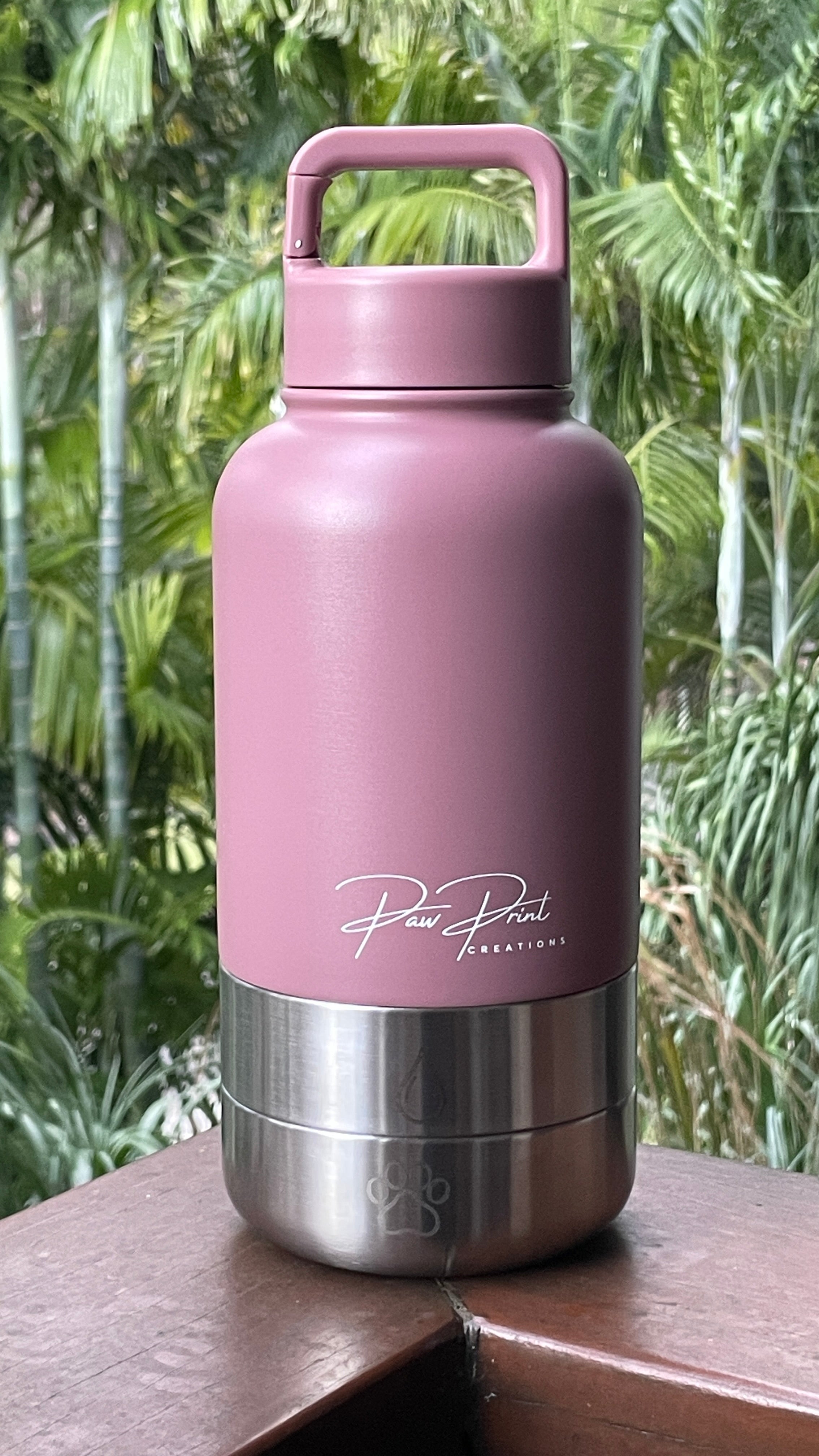 A dusty pink insulated water bottle with a stainless steel base, featuring the 'Paw Print Creations' logo, resting on a wooden ledge with a blurred tropical green background.